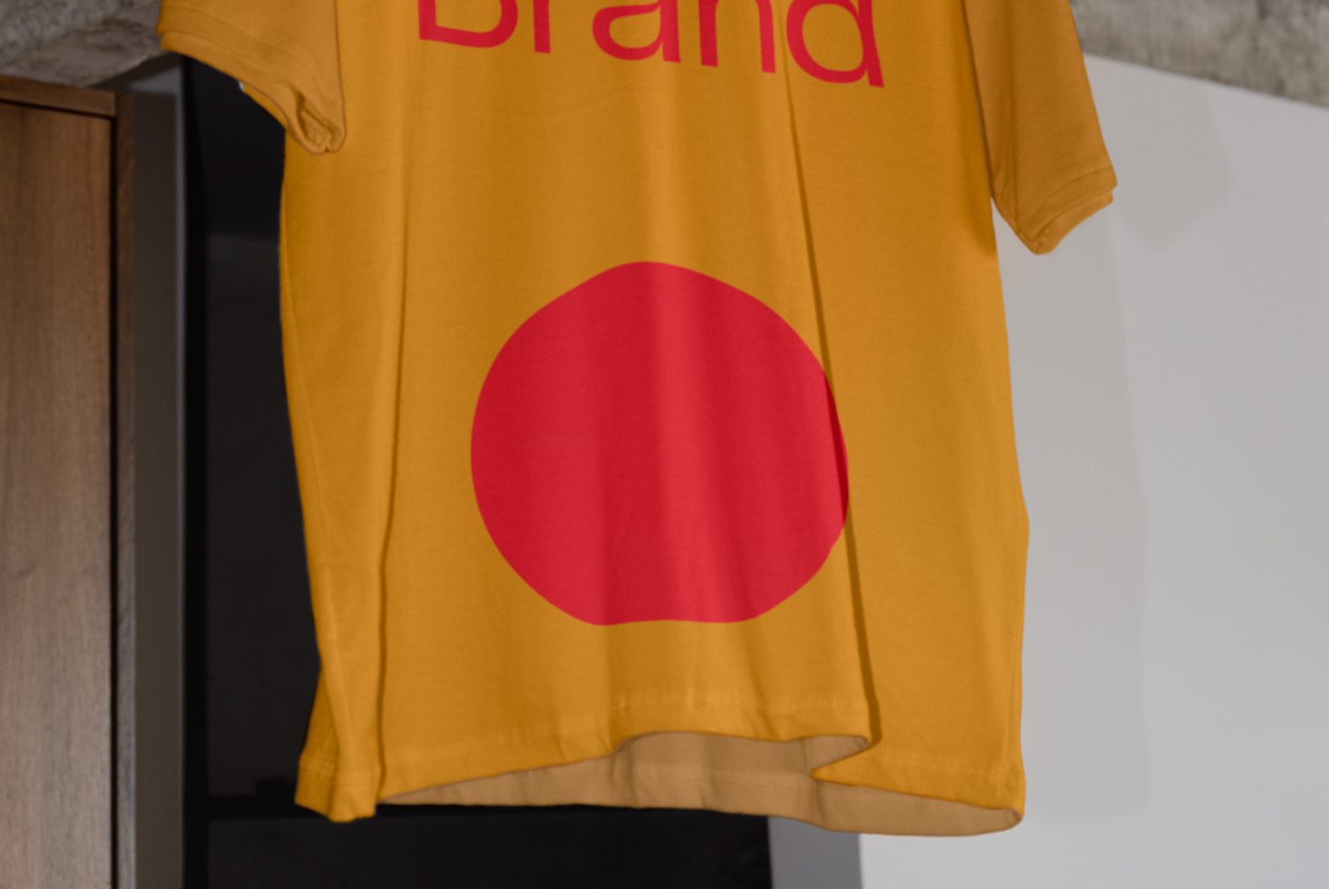 Yellow T-shirt mockup hanging on a wall featuring a red circle and brand text, ideal for fashion design presentations.